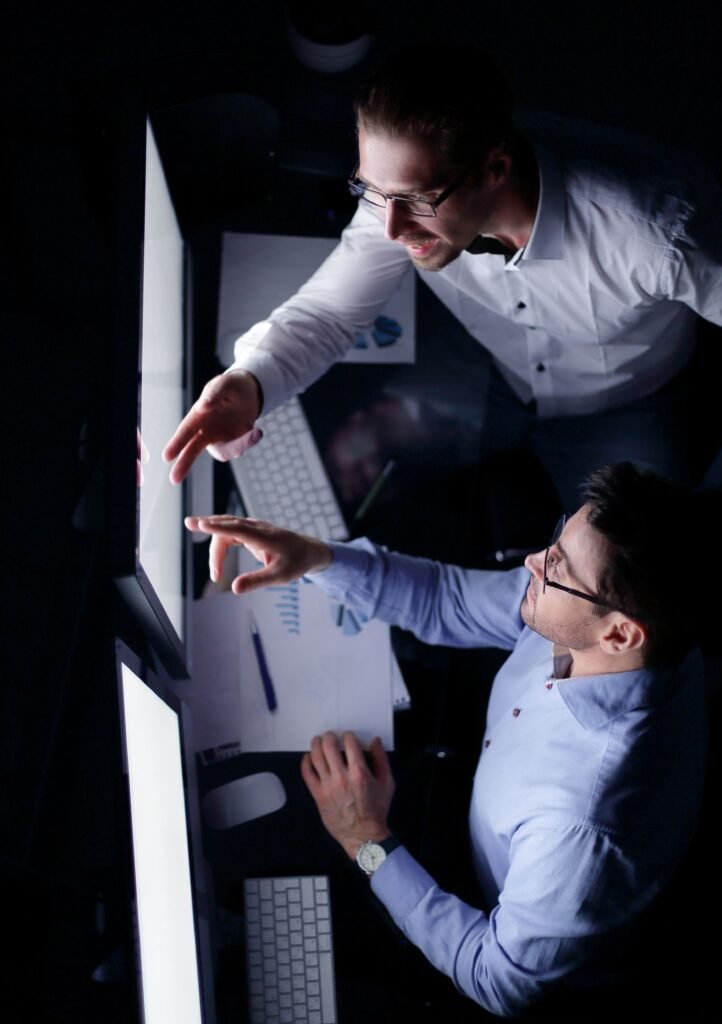 Vista de cima de dois colegas de trabalho discutindo informações na tela de um computador em um ambiente de escritório escuro. Um dos colegas aponta para a tela, enquanto o outro segue as instruções com o dedo, representando uma consultoria em tecnologia.