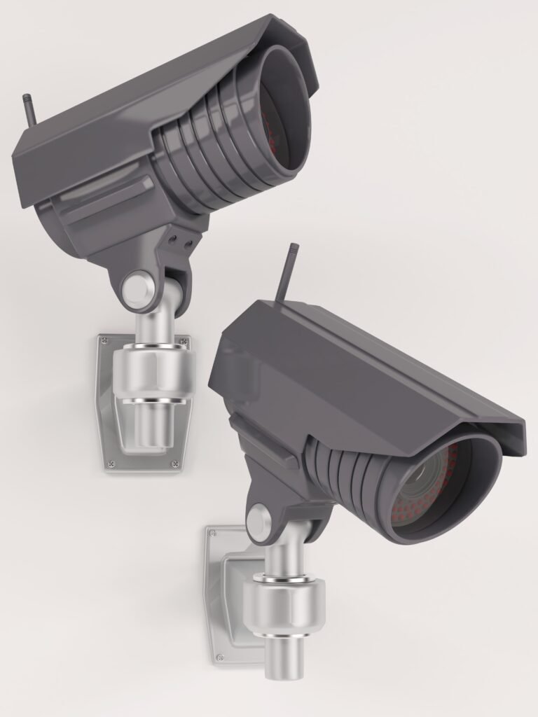 A imagem mostra duas câmeras de segurança modernas, de cor cinza, montadas em suportes articulados. Cada câmera é robusta, com um design que inclui uma lente grande e um revestimento protetor ao redor, sugerindo alta durabilidade e adequação para uso externo. Elas parecem ser modelos de alta definição, possivelmente com capacidades de visão noturna, como indicado pelos anéis vermelhos ao redor das lentes.