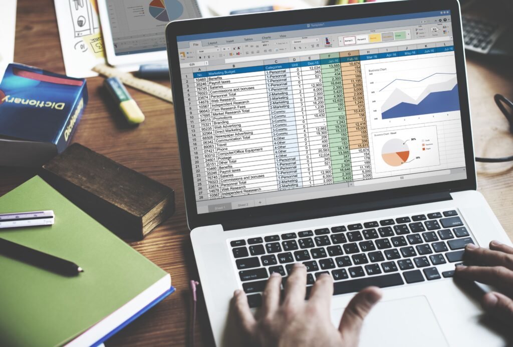 Uma pessoa trabalhando em um laptop com uma planilha de Excel aberta na tela. A planilha exibe dados financeiros organizados em colunas e linhas, com gráficos e tabelas visíveis. A mesa de trabalho tem vários itens, incluindo livros, cadernos e canetas.